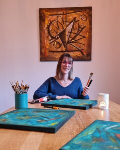 Elodie Moulin, au bout d'une table, avec 3 toiles en cours, des pinceaux et de la peinture